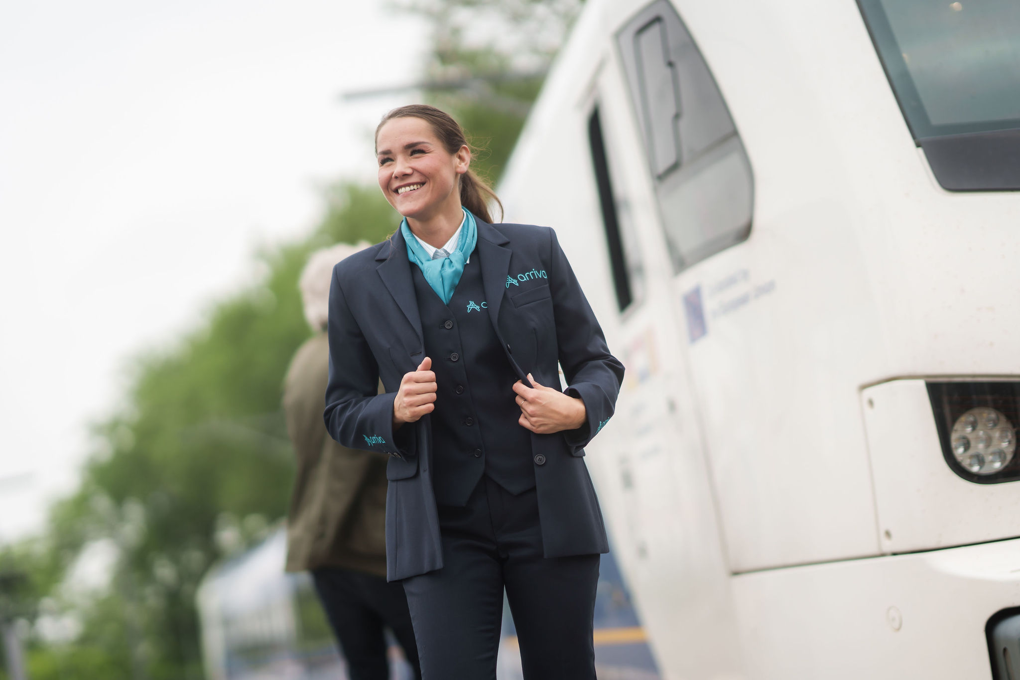 Een lachende machinist in Arriva-uniform staat buiten naast een trein, met bomen en een andere persoon op de achtergrond.
